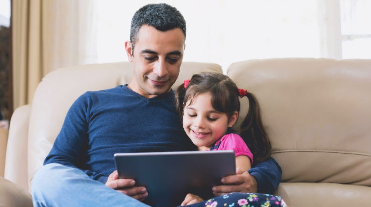 A father sits on the sofa with his young daughter. Both are immersed in the content of a laptop screen and the girl is smiling.