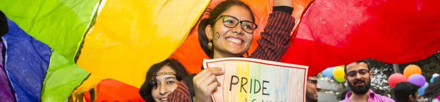 A group of young Asian women march in a Pride parade, their faces painted with rainbows.