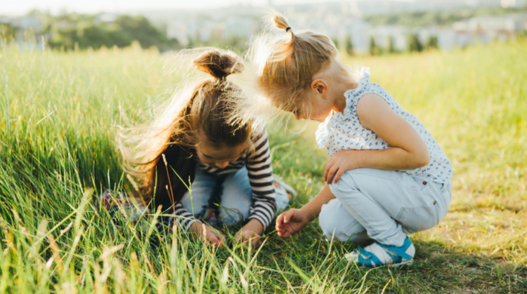 Two young girls in a field. They're crouching down, looking closely at something small hidden in the grass