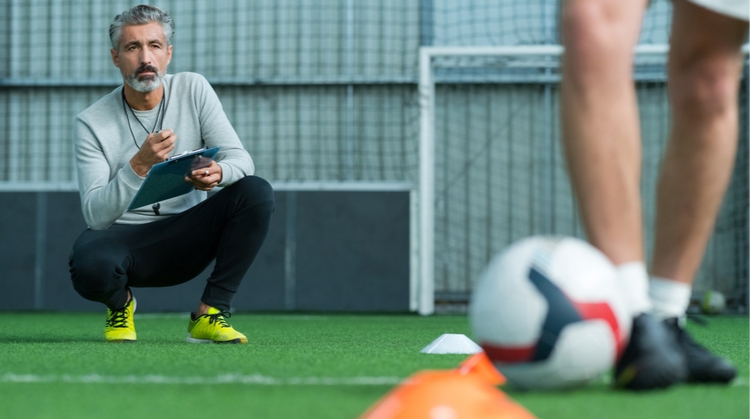 A football coach is crouched down with a stopwatch in one hand and a blue clipboard in the other. He is observing a football player who is kicking a ball around some cones. We can only see the player's legs and feet. 