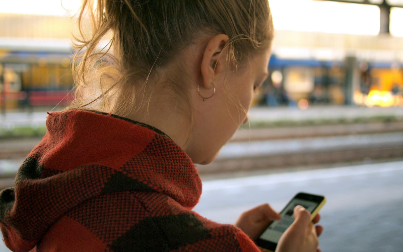 A blonde girl wearing a red hoodie and reading her mobile phone at a train station