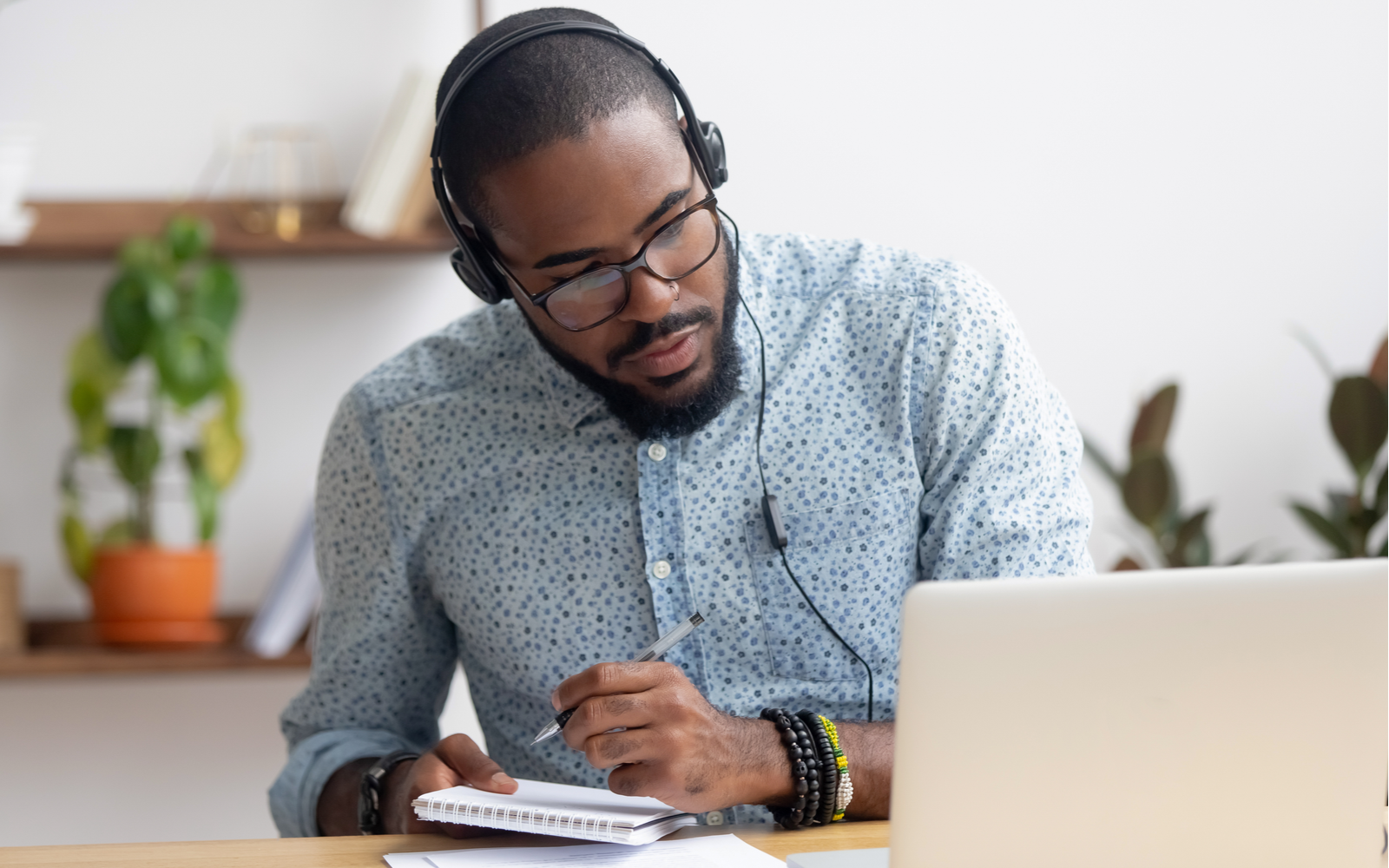 A man is sitting at a laptop computer, staring intently at the screen. He's wearing a headset, and is taking notes on a reporters notepad. 