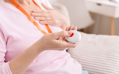 An elderly lady wearing a pink sweater is holding a remote personal alarm. It's around her neck with a lanyard and she's pressing a large red button. 