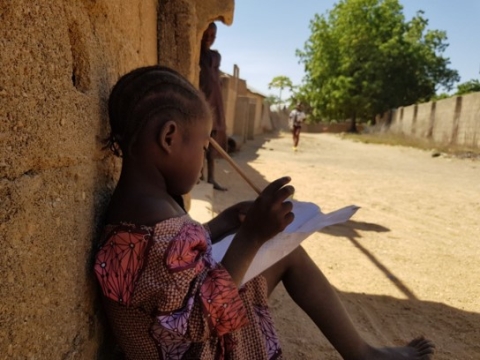 A photo of a young girl sat outside in Nigeria writing on a piece of paper