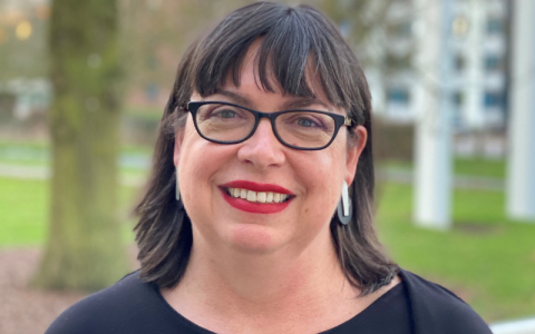 Portrait photograph of Professor Mimi Tatlow-Golden. She is wearing glasses and looking directly at the camera. Behind her you can see the OU buildings