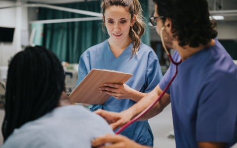 A student nurse takes notes on a clipboard while another nurse examines a patient in a hospital