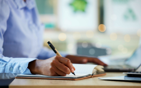 A woman writing in a notebook with a laptop beside her