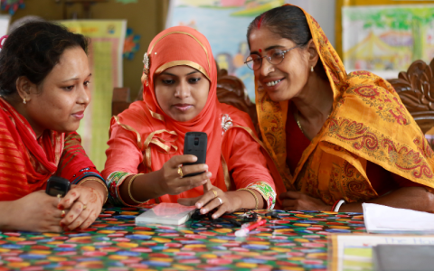 Three women sit at a table, they are all watching a mobile phone intently. 