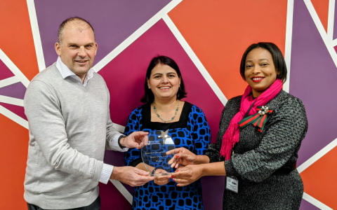 Three people holding a glass trophy are stood together in front of a colourful wall