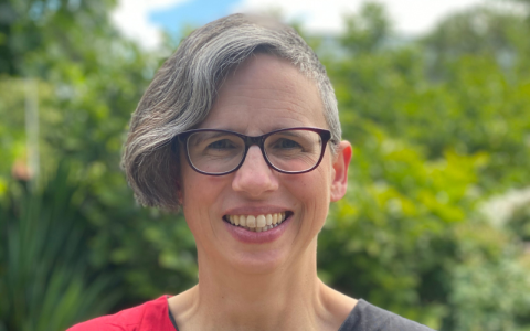 Portrait photograph of Professor Rebecca Jones. She is in an outdoor setting with greenery behind her. She is wearing glasses and a red and black top, and is smiling into the camera. 