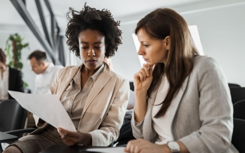 Two women sit side by side, they are look intently at a document. 