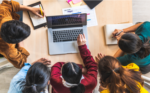 An aerial image of 5 people sitting around a table, sharing a computer. Some are taking notes and are all looking intently at the screen.