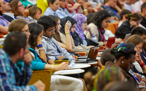 Young people from around the world sit in a large auditorium. They're looking towards a speaker who is off camera. 