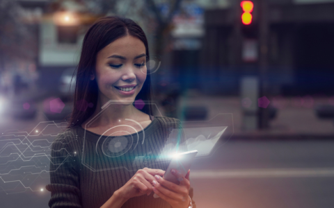 A young Asian woman is in the street, looking at her phone, as an augmented reality projection emerging from it..