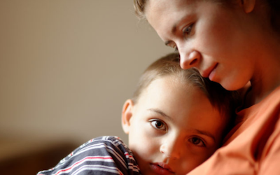A young boy sits on a woman's lap, nestled into her chest. He is staring sadly into the camera, she has her cheek resting on the top of his head.