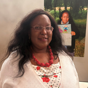 Photo of Donna Harrison wearing a red necklace and stood in front of a childhood photo of herself