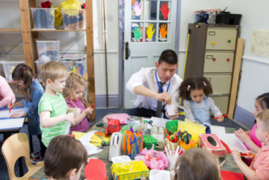 A teacher and diverse group of students play with coloured paper and stationery around a table