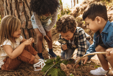 A diverse group of children play in the wild - one is looking at leaves through a magnifying glass