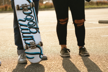 A feet-up shot of two teenagers. One wears jeans ripped at the knees, one carries a skateboard