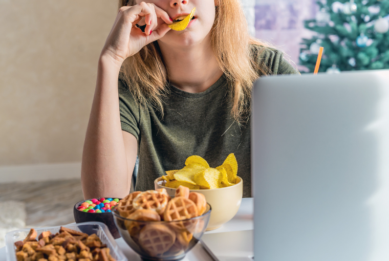 Teenager using a computer eats crisps from a selection of sugary and salty snacks on the table