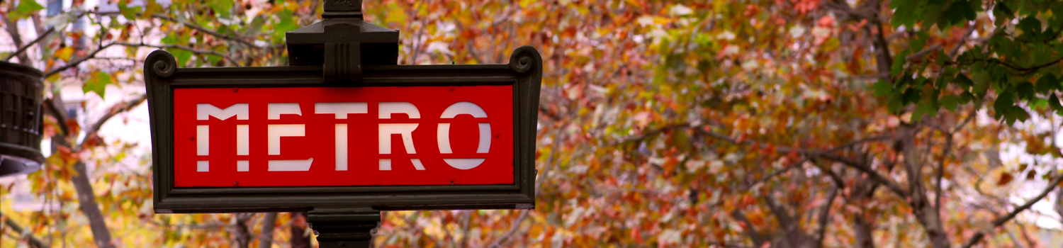 An old-fashioned iron sign for the Metro system in Paris sits against a backdrop of leafy red and green trees and a grey Autumn sky.