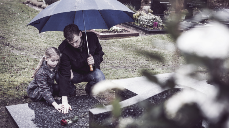 A father and daughter at a graveyard on a grey, rainy day visiting the grave of their wife/mother. They are both kneeling at the foot of a grave, touching the wet earth; the father holds a large blue umbrella over them both.