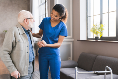 A nurse in scrubs helps an elderly man walk with a zimmer frame