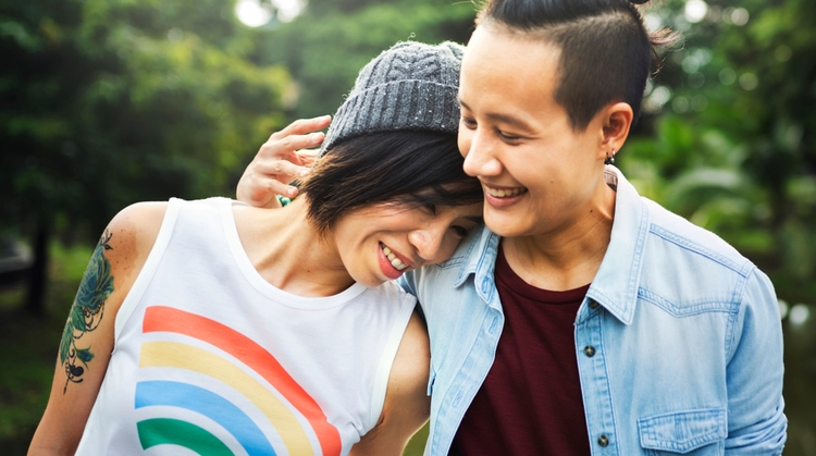 A happy LGBT couple embracing each other and smiling. The lady on the left is wearing a white sleeveless t-shirt with a rainbow image on it