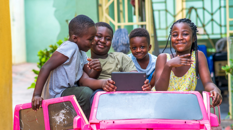 Four Ghanaian children (three boys and one girl) are sitting in a large pink play car. They're smiling and one of the boys is holding a tablet.