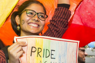 A young woman with glasses smiles broadly and holds a poster with the word 'PRIDE'