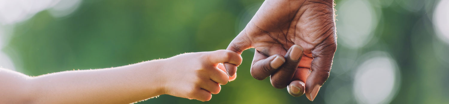 Shot of a young child's arm holding on to an elderly person's hand by the little finger. The older arm wears a brown leather watch with a gold buckle. The background is distorted greenery,