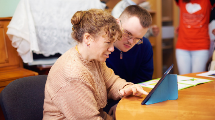 two friends, one male, one female, with a learning disability watch a digital tablet together, whilst seated at a table. 