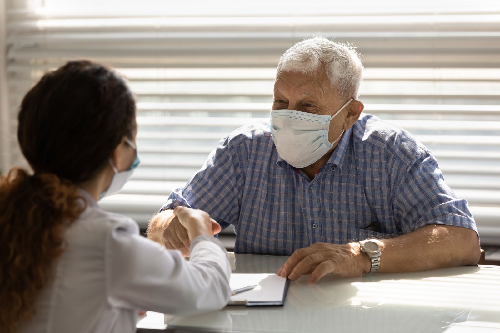 Smiling senior male patient in medical facial mask shake hand close health insurance deal with female doctor. Happy mature man client in facemask handshake woman GP at meeting in hospital.