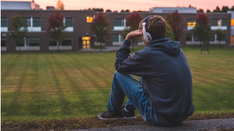 teenage boy sits overlooking a field. He's wearing a hooded sweatshirt and facing away from the camera. He's wearing headphones and looking at his phone