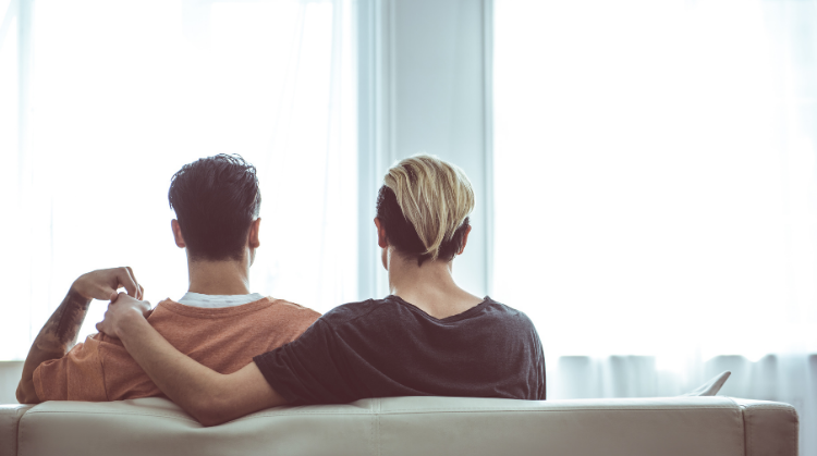 Back view portrait of young man with dyed hair hugging boyfriend while he touching his hand. They are resting on sofa in front of windows.