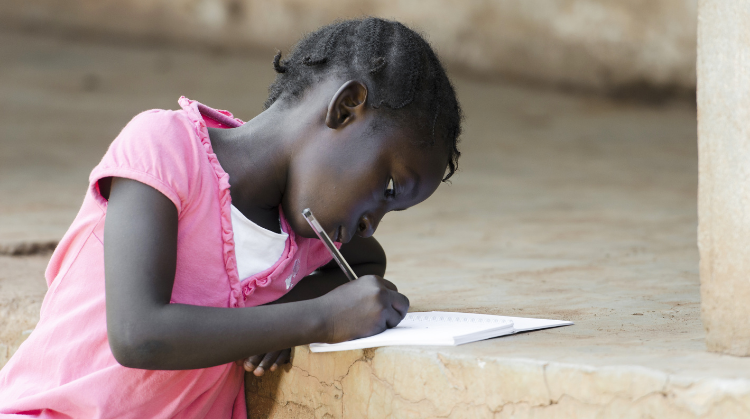 A young, black African girl sits on a step outside writing in a note book.