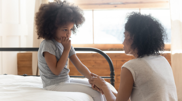 African american mother comforts her young daughter. She is holding her hand and kneeling down in front of her as she sits on a bed, looking sad and worried.