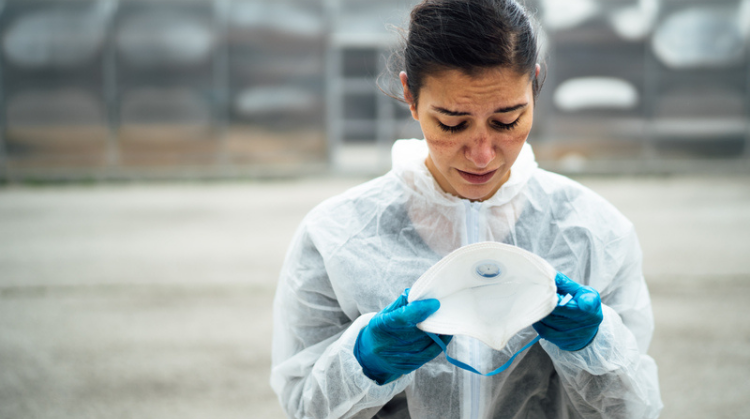 An exhausted female doctor/nurse in coronavirus protective gear stands outside clutching their N95 mask. They are looking down tearfully.