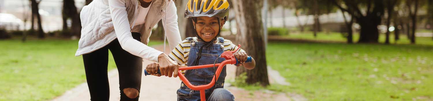 A mother supports a young child learning to ride a bike. The child is smiling widely and wobbling slightly on the bike,