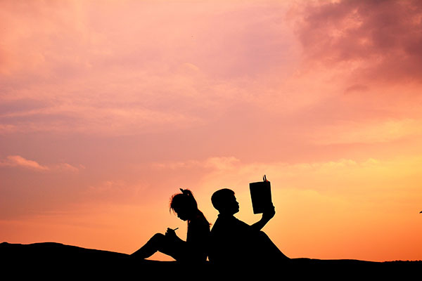 silhouette style picture that shows two people sitting back to back with each other - one is reading a book, another one is writing something with a pen.