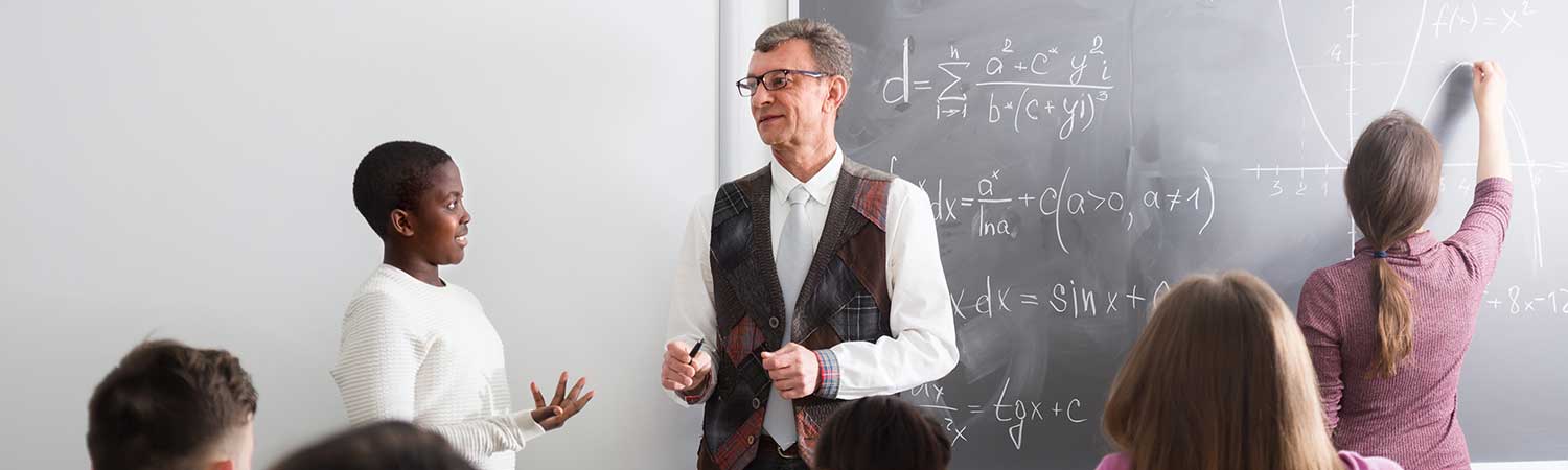 group photo of a classroom with a student talking to his teacher in front of a class while another students writes on a chalkboard