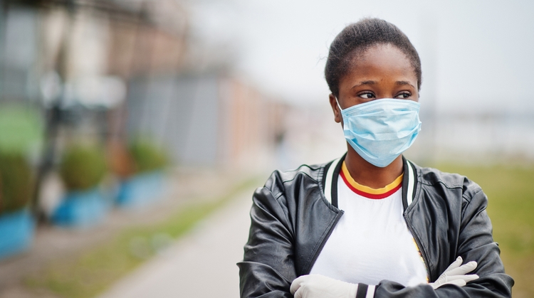 Black teen girl, wearing a blue surgical mask, staring into the distance with her arms folded