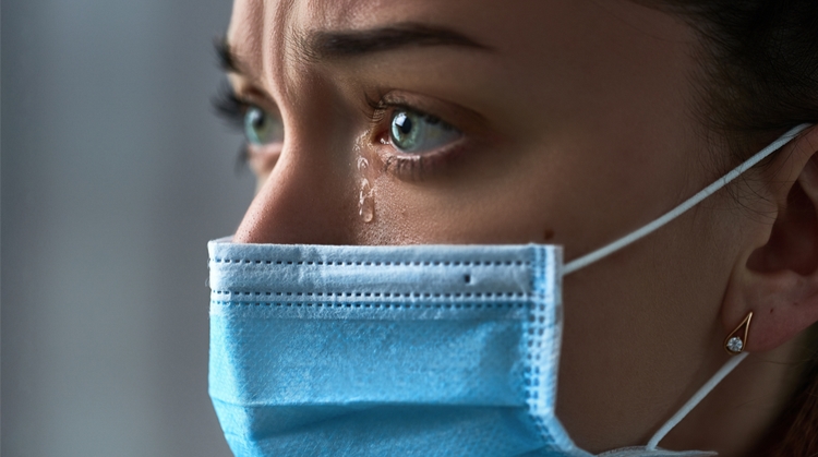 Close up of a woman's face. She is wearing a surgical mask and staring into the distance. A tear is running down her face. 
