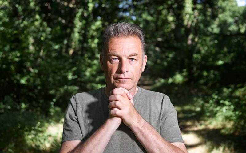 Chris Packham stares into the camera. His hands are clasped beneath his chin, he is in a wooded area.