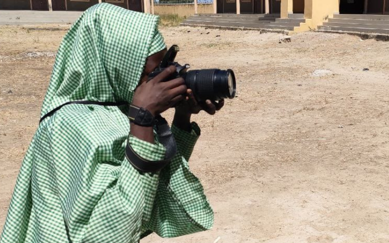 A young girl from the Lake Chad area is taking a photograph. Her face is obscured by the camera and she's wearing a green and white hijab