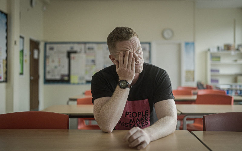 Rapper Darren McGarvey sits at a school desk. He is resting his head in his hand, covering part of his face. His t-shirt reads: people make mistakes. 