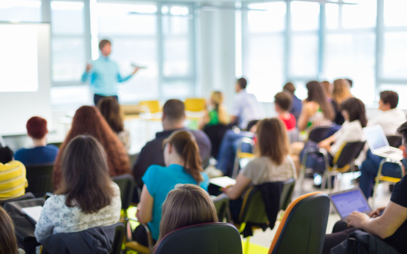 a man stands at the front of a classroom of older students, who have their backs to the camera. All the people are out of focus