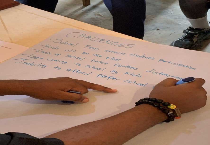 Several participants stand around a display board covered with photographs and captions, reviewing and discussing the visual materials during a community workshop or project presentation.