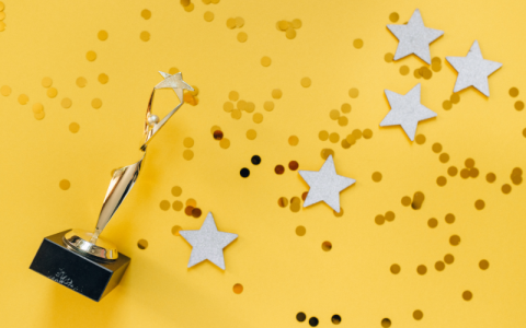 A trophy in the shape of a person with their arms aloft is lying on a yellow background which is covered in stars and confetti. 