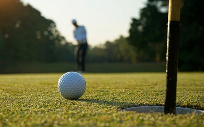 close up of a golf ball, on the green, inches away from the hole. The golfer can be seen, out of focus, in the distance.